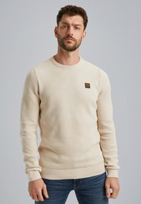 Pull texturé beige avec un col rond, des poignets et un ourlet côtes, arborant un petit écusson de logo sombre sur la poitrine gauche. Porté avec un jean bleu.