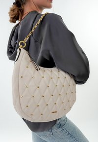 Beige quiltet håndtaske med guldnitter, en guld kædestropp og glat lædertekstur. Moderne hobo-form med en karakteristisk logo-detalje.