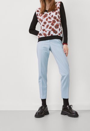 Femme portant un pantalon bleu clair, des chaussures noires épaisses, un haut à manches longues noir et un gilet blanc avec un motif floral marron, debout contre un mur blanc.