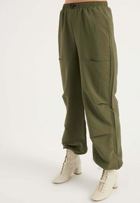 Olijfgroene, lichte broek met een elastische tailleband en twee grote zijzakken, gecombineerd met crèmekleurige, puntige hoge hakken.