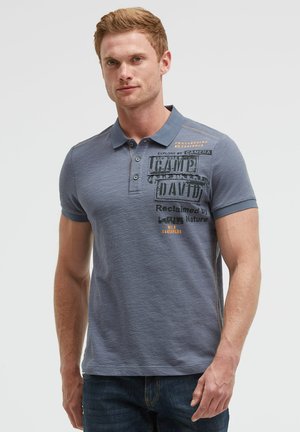 Mann mit kurzen roten Haaren, der ein blaues Polohemd mit Text und dunkle Jeans trägt, steht vor einem einfarbigen Hintergrund.
