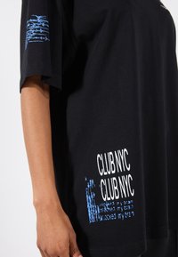 Black cotton t-shirt with blue graphic text on the sleeve and front; loose fit with short sleeves and a round neckline.