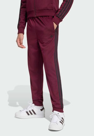Persona que lleva pantalones deportivos y chaqueta burdeos con rayas negras, combinados con zapatillas blancas con rayas negras.