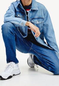 Persona agachada con chaqueta de mezclilla azul claro, jeans azul oscuro, camisa de rayas y zapatillas blancas sobre un suelo blanco.