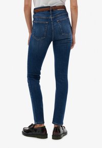 Jeans de denim azul oscuro, ajustados y de cintura alta, con bolsillos traseros y un cinturón trenzado marrón, combinados con zapatos negros.