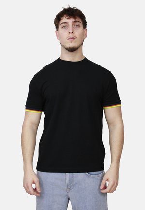 Uomo che indossa una maglietta nera con bordi delle maniche in rosso, arancione e giallo, e jeans azzurri, in piedi davanti a uno sfondo bianco.