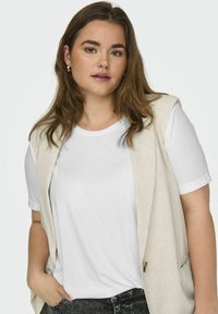 Un gilet sans manches beige clair porté sur un t-shirt blanc, associé à un jean noir. Le gilet présente une coupe décontractée et un design minimaliste.