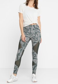 Legginsy w zielono-białym geometrycznym wzorze, z przezroczystymi panelami. Połączone z luźną białą koszulą i białymi butami sportowymi.