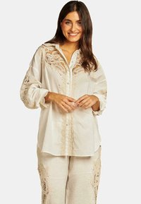 Blusa in lino beige con dettagli in pizzo intricati, patta dei bottoni e maniche lunghe e svolazzanti. Tessuto testurizzato con accenti scallop.