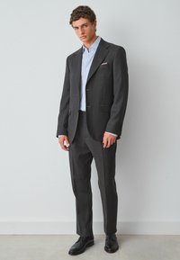 Grau Anzug mit maßgeschneiderter Passform, subtiler Textur und aufgesetzten Revers. Getragen mit einem hellblauen gestreiften Hemd und schwarzen Schuhen.