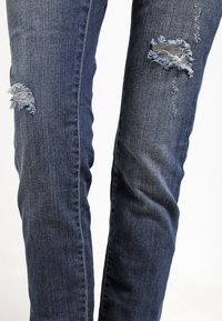 Jambes portant un jean en denim bleu foncé avec des déchirures effilochées sur le haut des cuisses sur un fond uni.