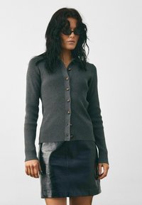 Szürke bordázott kardigán barna gombokkal, fekete fényes bőr szoknyával párosítva. A textúrált anyag és a testes szabás kiemeli az öltözéket.