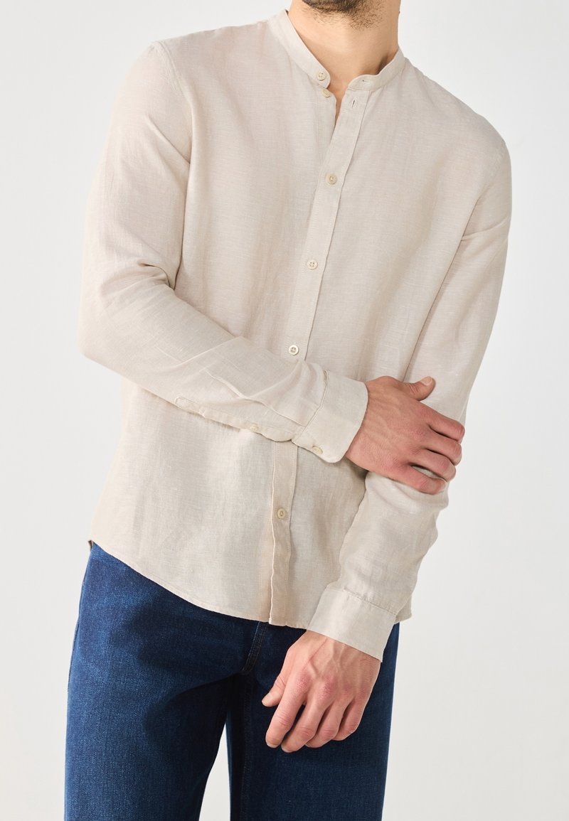 Lichtbeige linnen overhemd met knopen, mandarijnkraag en lange mouwen, gecombineerd met donkerblauwe denim jeans. Subtiele textuur zichtbaar.