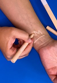 Crayon de maquillage beige clair dans la main, en application sur la peau. Échantillon sur le bras montrant une forme de couronne irrégulière. Emballage plat à proximité sur fond bleu.