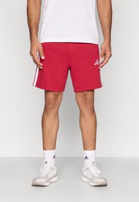 Raudoni sportiniai šortai iš minkšto medvilnės mišinio, su baltais šonais dryžiais ir „Adidas“ logotipu ant kairės kojos. Klasikinis dizainas ir prigludimas.