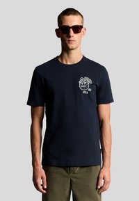 Marineblå bomulds t-shirt med et hvidt grafik af en løve og tekst på venstre bryst. Korte ærmer og crew neck-design.