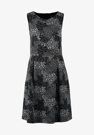 Femme portant une robe sans manches noir et gris à imprimé animal, collants noirs, bottines, tenant une pochette beige sur le côté.
