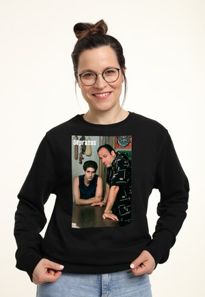 Schwarzes Sweatshirt mit einem Fotodruck von zwei Männern, die sich an einen Tisch lehnen; einer trägt ein blaues Tanktop, der andere ein gemustertes schwarzes Hemd.