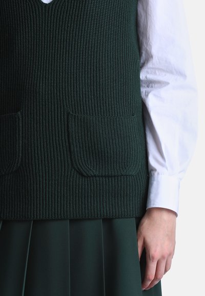 Main portant une chemise blanche à manches longues et un gilet en tricot côtelé vert foncé avec deux poches avant sur une jupe plissée vert foncé.
