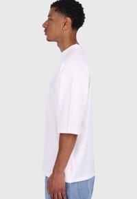 Hvid bomulds t-shirt med korte ærmer, rund hals og sideslids. Har et subtilt logo på ærmet. Modellen står sidelæns.