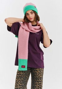 Růžová a zelená pletená čepice a odpovídající šála. Šála má ribbovaný zelený akcent. Model nosí fialové tričko a kalhoty s leopardím vzorem.