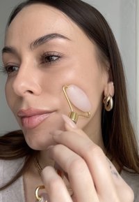 Rouleau facial en quartz rose avec un manche en or, utilisé sur la peau. La main de l'utilisateur présente de longs ongles neutres et des bijoux discrets. La peau semble lisse et radieuse.