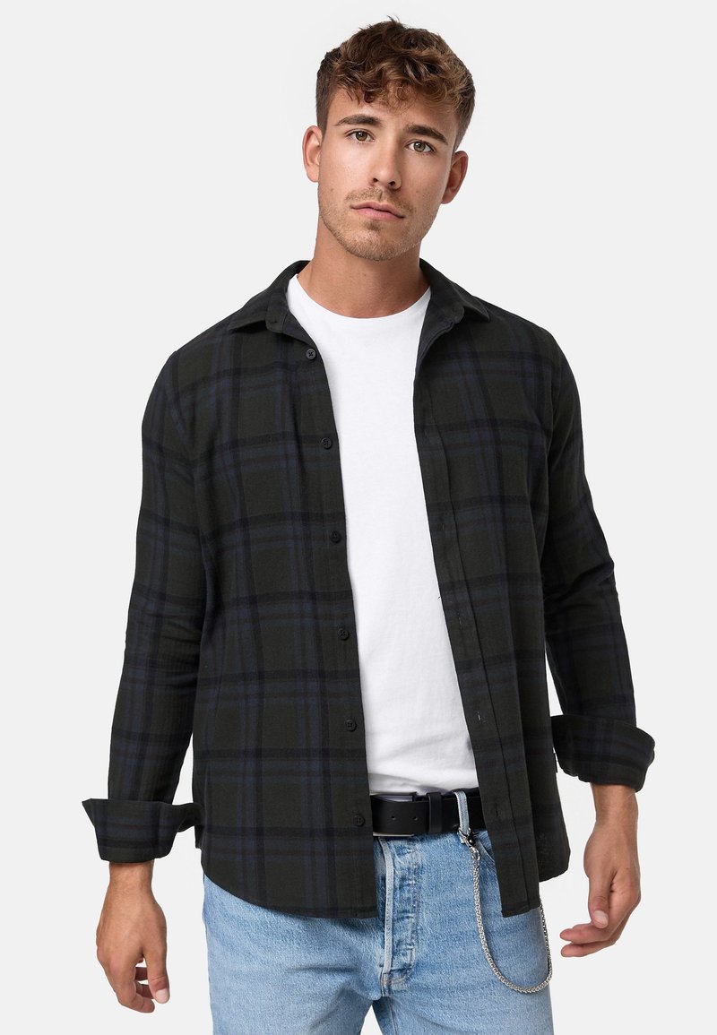 Camisa de franela de cuadros negra y verde con diseño de botones, mangas largas y dobladillo curvado, combinada con una camiseta blanca de cuello redondo y jeans de mezclilla.