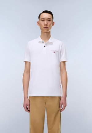 Ung mand stående iført en hvid poloshirt med logo og beige bukser mod en ensartet lys baggrund.