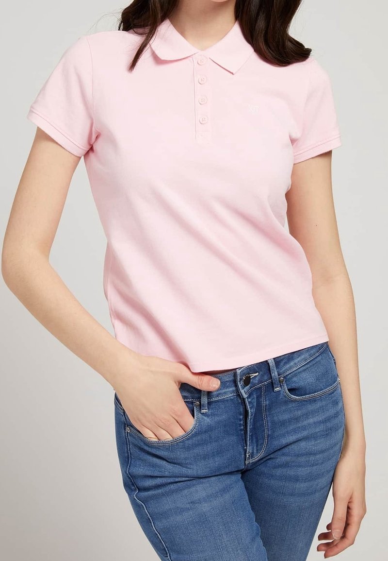Polo en tejido rosa suave, con cuello, mangas cortas y tapeta de cinco botones. Combinado con unos jeans de denim azul ajustados.