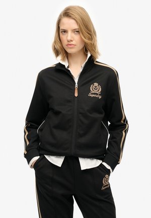 Veste de survêtement zippée noire avec des accents dorés et le logo "Superdry" brodé, associée à un pantalon noir. Présente des poches latérales et un col.