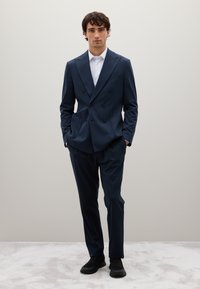 Marineblauer zweireihiger Anzug mit tailliertem Schnitt, ausgestattet mit einem Revers, zwei Fronttaschen und eleganten Schuhen. Glatte Stoffbeschaffenheit.