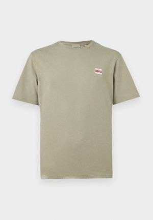 T-shirt vert clair à manches courtes et col rond avec un petit patch logo rectangulaire rouge et blanc sur le côté gauche de la poitrine.