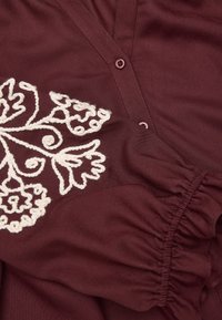 Burgundinyn blouse valgete lillemustriga ühel küljel, nööbiga kinnitamine ja rikutud varrukade detailid mugava istuvuse jaoks. Pehme kangastekstuuri.