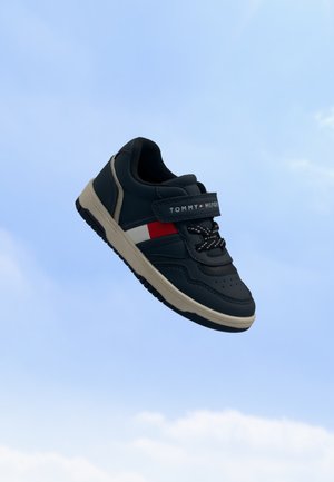 Baskets enfant noires et beiges avec le logo Tommy Hilfiger, bandes latérales rouges et blanches, et une bride velcro, sur fond de ciel bleu.