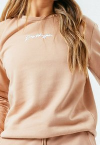 Světle růžová mikina vyrobená z jemné tkaniny, se zaobleným výstřihem a bílým výšivkou "Just Hype" na hrudi.