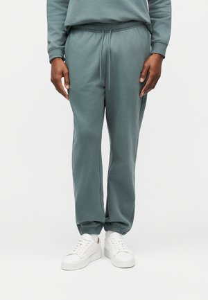 Osoba nosząca spodnie dresowe w kolorze łupkowo-zielonym z elastycznymi ściągaczami i paskiem ściągającym w talii, w połączeniu z białymi, niskimi trampkami, stojąca na jednolitym tle.