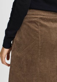 Jupe en velours côtelé marron avec des côtes verticales, présentant une surface lisse et texturée et une couture arrière nette. Main reposant sur le côté.