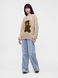 Femme portant un pull beige moucheté avec un motif d'ours en peluche marron et un jean large bleu clair, debout contre un fond blanc uni.