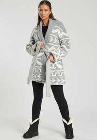Grå mönstrad omslagsjacka med vita geometriska mönster, stickad textur, breda slag och knytliv, kombinerad med svarta leggings och stövlar.