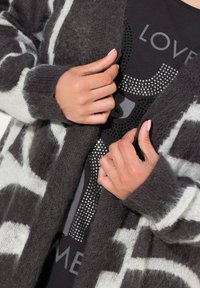 Übergroßer Cardigan in Dunkelgrau mit weißen Streifen und weicher Textur, getragen über einem schwarzen Top mit metallischen Verzierungen.