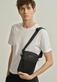 Czarna torebka typu crossbody z fakturowanym panelem, zamknięciem na zamek i solidnym, regulowanym paskiem. Materiał wydaje się trwały i lekki.