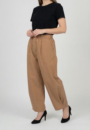 Donna che indossa una maglietta nera a maniche corte, pantaloni larghi color sabbia con vita elastica e scarpe nere con tacco alto, in piedi su sfondo bianco.