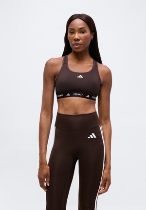 Nő barna Adidas Techfit sportmelltartóban és hozzáillő, fehér csíkos magas derekú leggingsben, áll egy egyszerű, világosszürke háttér előtt.