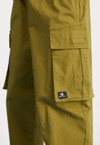 Spodnie typu cargo w oliwkowej zieleni, wykonane z teksturowanej tkaniny, z bocznymi kieszeniami oraz małą czarno-białą naszywką z logo Champion na kieszeni.