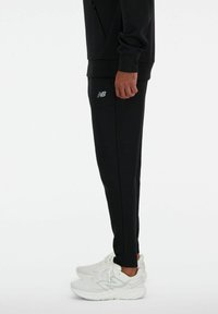 Pantaloni jogger neri con una vestibilità slim, dotati di un discreto logo New Balance sulla coscia e abbinati a scarpe da ginnastica bianche.