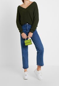 Olivgrön stickad tröja med V-ringning, kombinerad med höga blå denimjeans och vita sneakers. Håller en liten limegrön handväska.