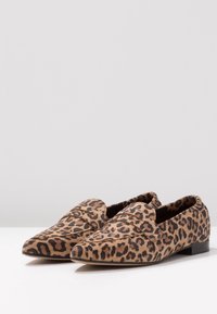 Une paire de mocassins à bout pointu avec un motif léopard marron et noir sur une surface blanche.