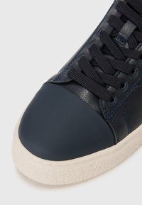 Marineblå lærersneakers med en mørk gummitåhætte, teksturert overflate og matchende snøringer. Lysfarget, teksturert gummisåle.