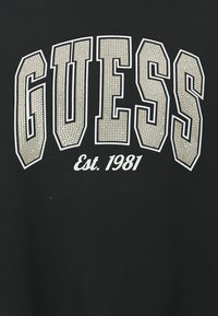 T-shirt nera con testo audace e sovradimensionato in bianco e argento "GUESS" con un motivo a pois e "Est. 1981" in testo bianco più piccolo sottostante.