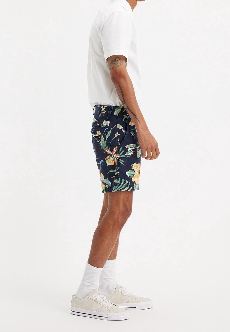 Homme portant une chemise blanche à manches courtes, un short bleu marine à motif floral, des chaussettes blanches et des baskets beiges, debout de profil.